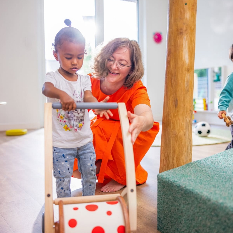 Pädagogin spielt hockend mit stehendem Kleinkind an einem Rollator